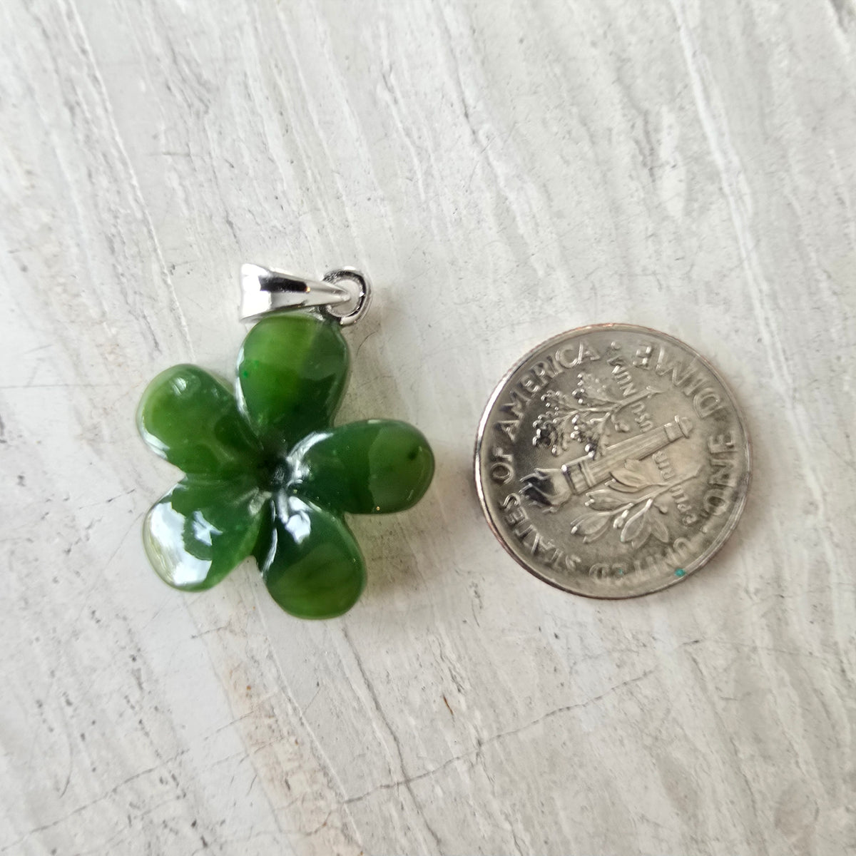 A green five-petal flower pendant made of polished stone or glass with a silver bail, shown next to a US dime coin for scale on a light wood surface.