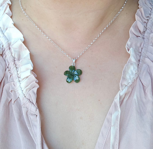 A silver chain necklace with a green five-petal flower pendant, worn by a person in a light pink ruffled blouse.