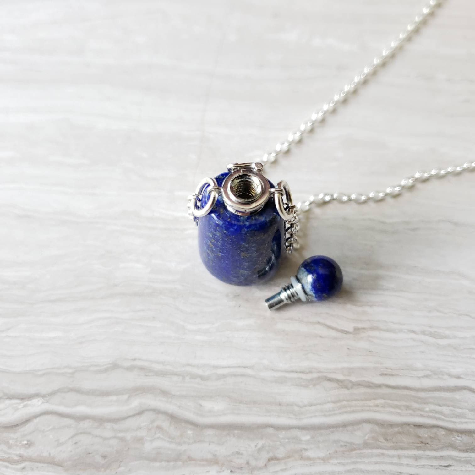A lapis lazuli bottle pendant necklace with a silver-colored cap and a chain, displayed against a textured background.