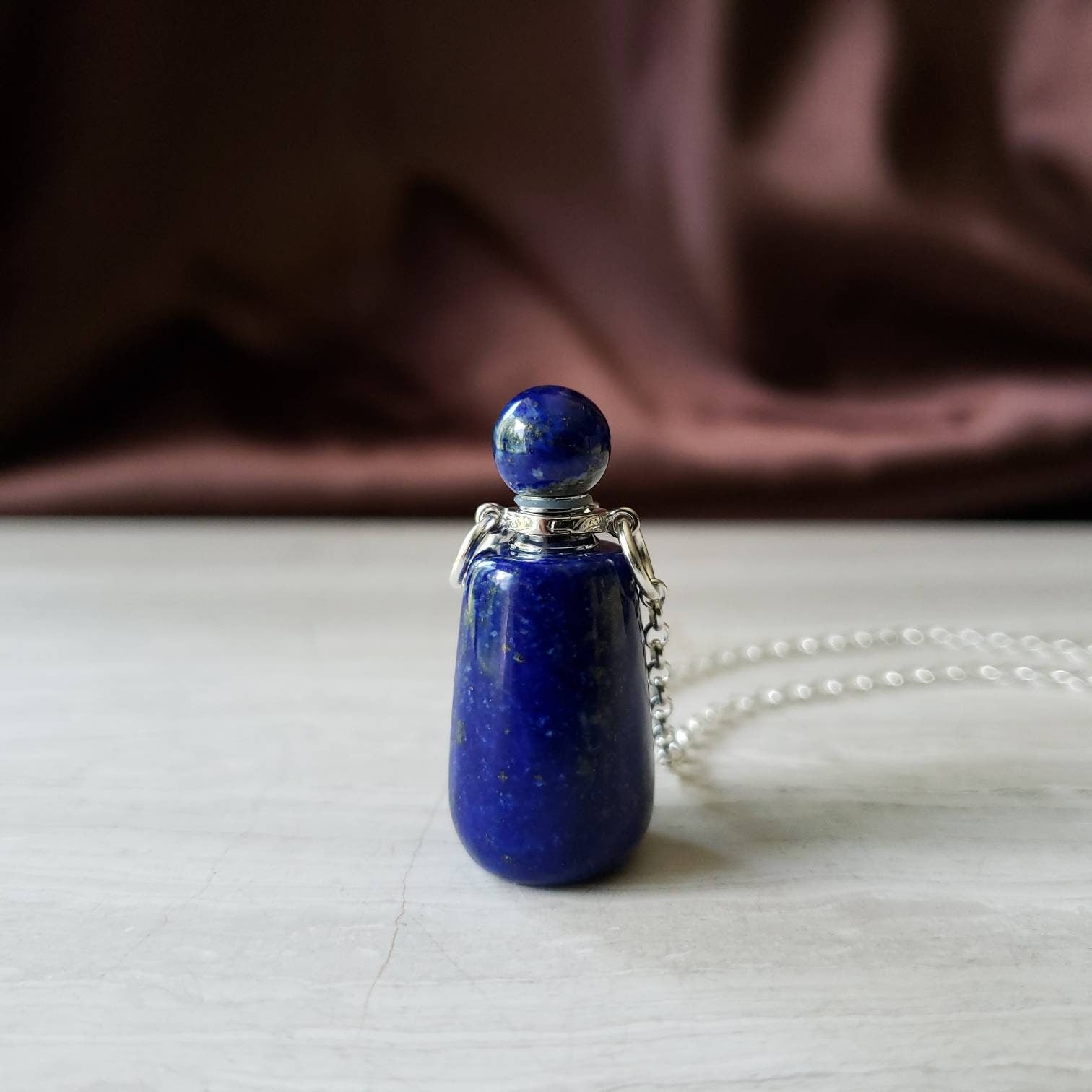 A lapis lazuli bottle pendant necklace with a silver-colored cap and a chain, displayed against a textured background.