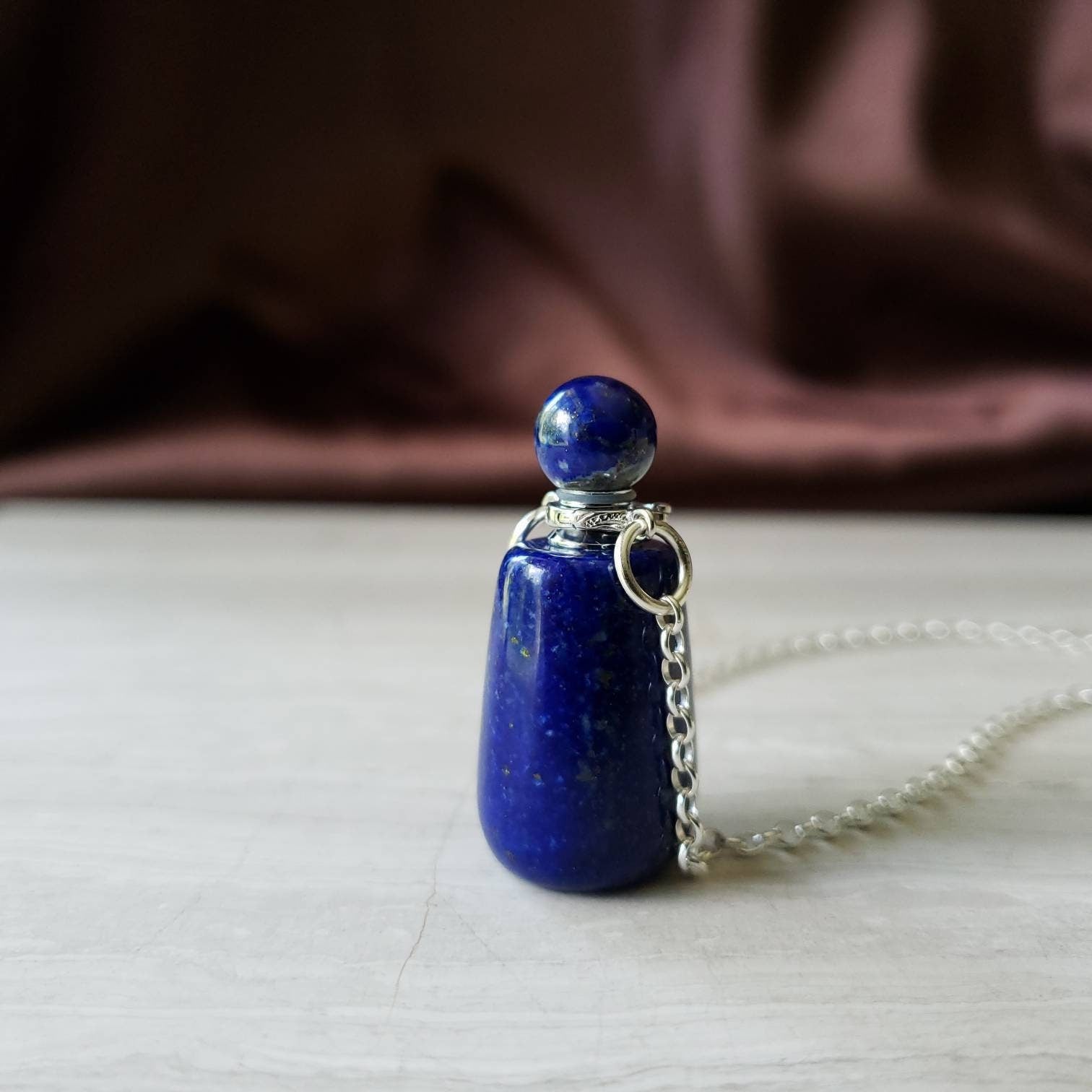 A lapis lazuli bottle pendant necklace with a silver-colored cap and a chain, displayed against a textured background.