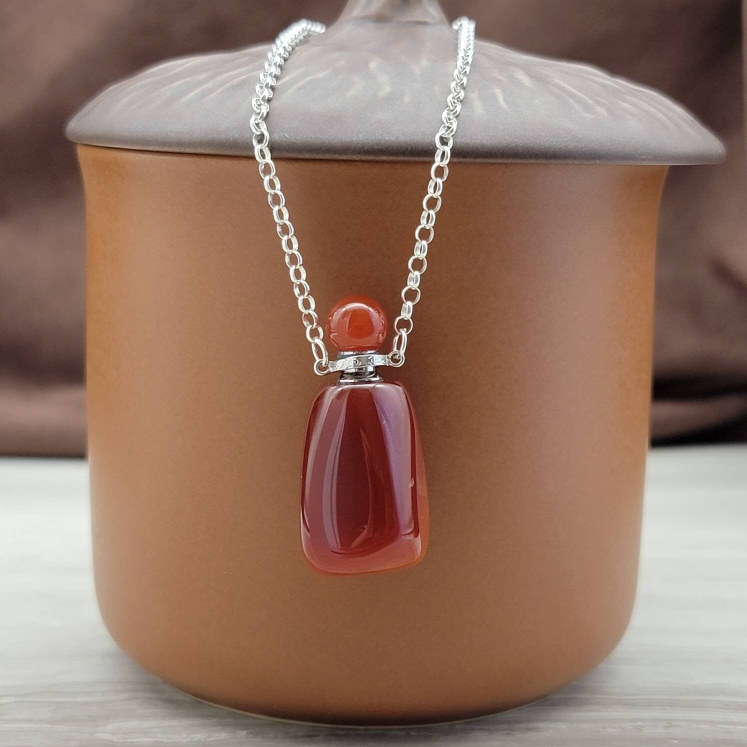A carnelian gemstone bottle necklace with a silver chain and a glass bottle inside a brown ceramic container.