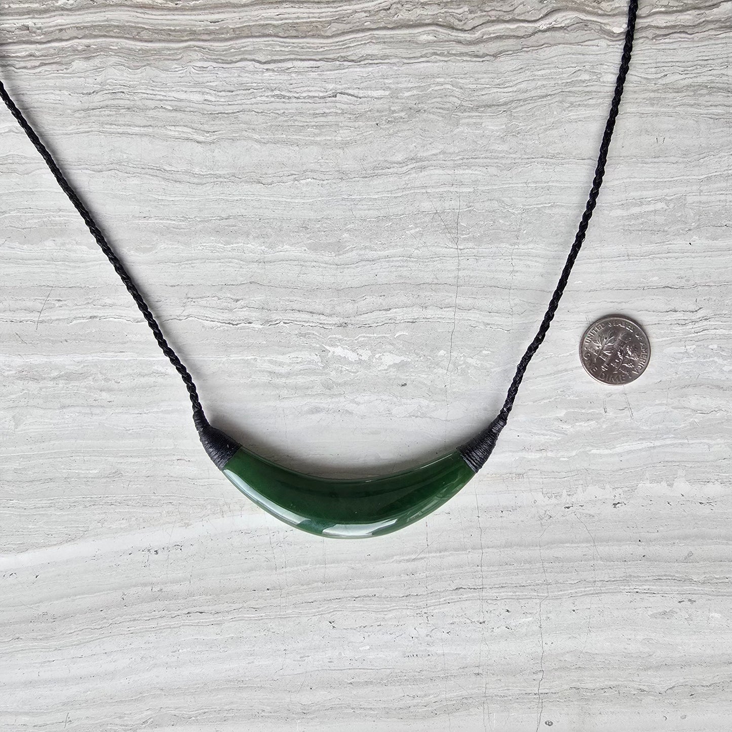 A green curved pendant necklace on a black braided cord, displayed on a light gray stone surface with a coin for scale.