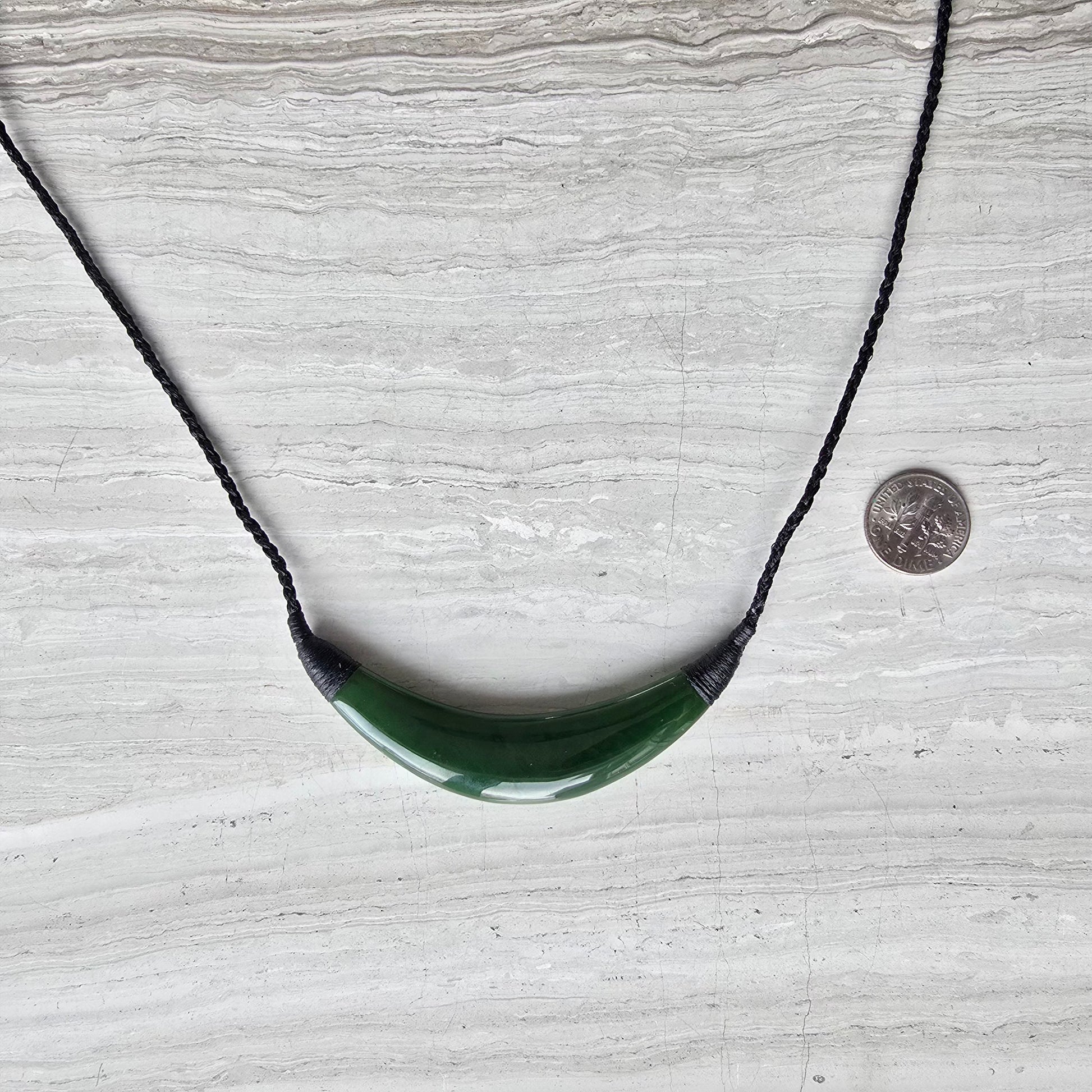 A green curved pendant necklace on a black braided cord, displayed on a light gray stone surface with a coin for scale.