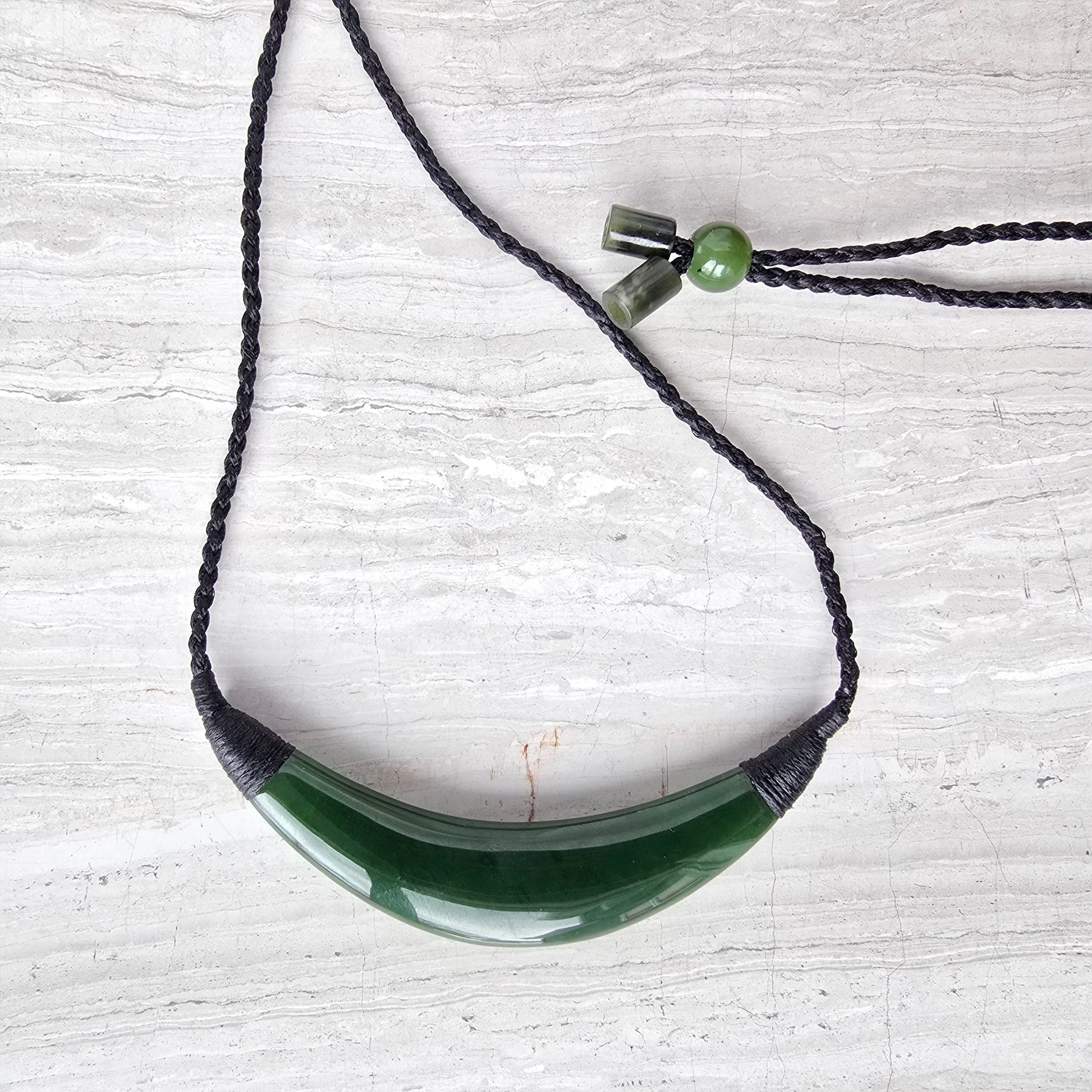 A green jade pendant necklace with a dark braided cord, displayed on a light gray wood-textured background.