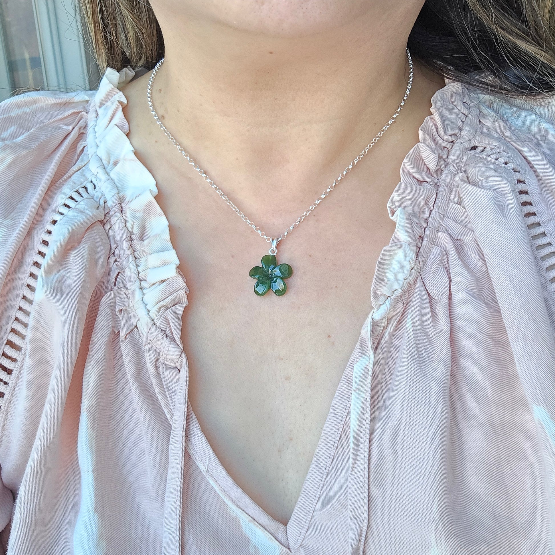 A close-up of a person wearing a silver chain necklace with a green four-petal flower pendant, paired with a light pink blouse.