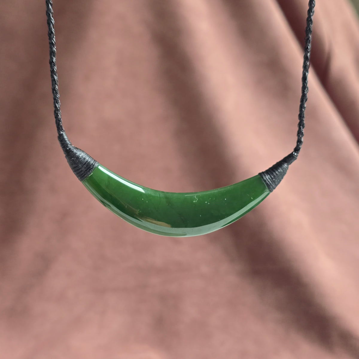 A green curved pendant necklace with a dark gray braided cord, shown against a soft-focus dusty rose background.