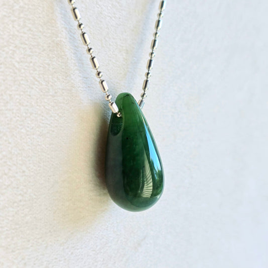 A polished dark green teardrop pendant necklace on a silver beaded chain, displayed against a light beige textured background.