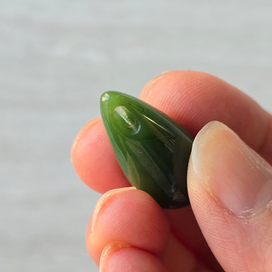 A polished green jade teardrop pendnt bead held between fingers against a soft light gray background.